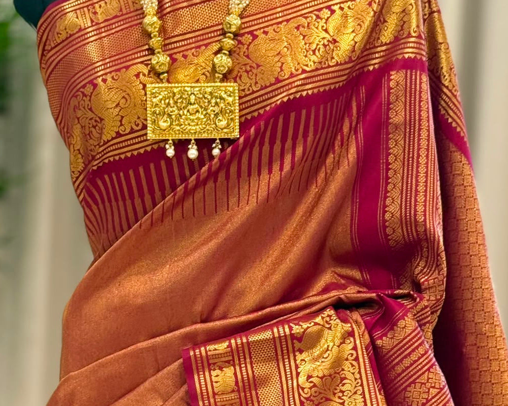 A close-up of a draped orange and gold zari silk saree with a deep magenta border, displayed on a black mannequin torso adorned with a gold necklace.