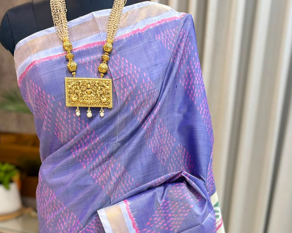 A purple and pink patterned silk saree draped on a black mannequin, styled with a gold beaded necklace, in an indoor setting with beige curtains in the background.